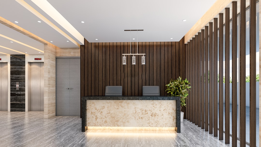 The image is a frontal view of a luxurious, modern reception area within a building lobby. A prominent reception desk, with a polished black granite or stone top and a glowing, translucent marble base, stands in the center. Behind the desk, a feature wall is composed of dark, vertical wooden slats. To the left, a set of elevators with brushed metal doors is visible, surrounded by walls with a mix of light-colored stone and a subtle black and white marble pattern. A potted plant sits to the right of the desk, and a contemporary light fixture hangs from the ceiling above it. The floor is a gloss